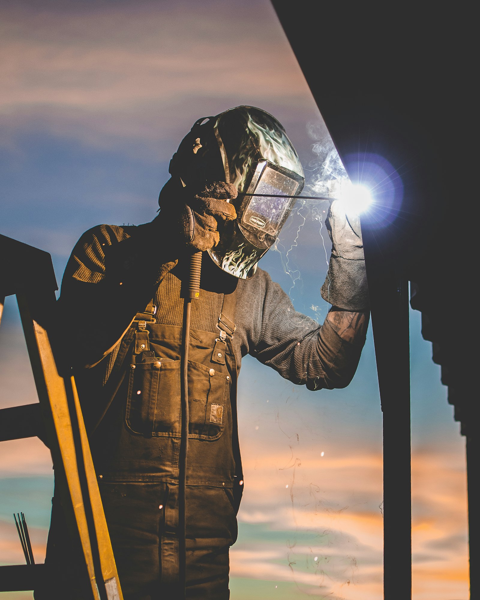 A welder working from a ladder on an outdoor structure, silhouetted against the sky